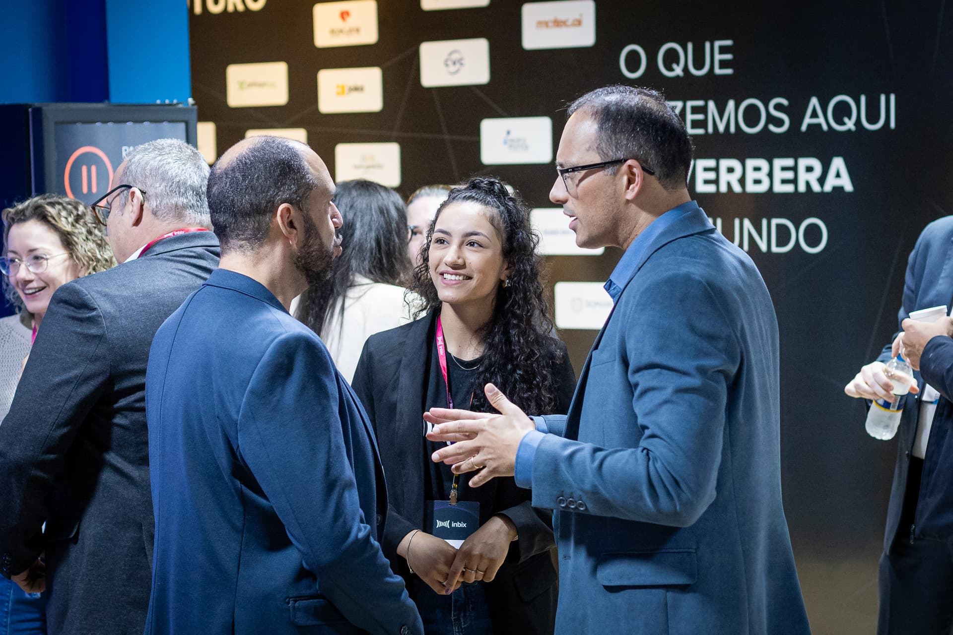 Profissionais conversando em evento diante do painel O que fazemos aqui reverbera no mundo