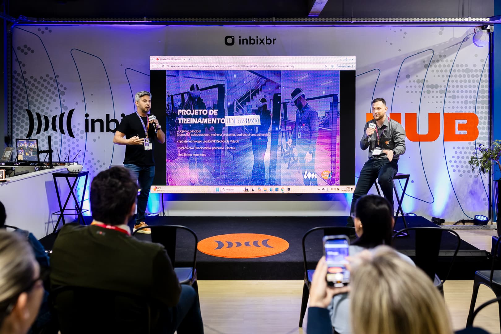 Dois apresentadores no palco com o slide de um projeto de treinamento